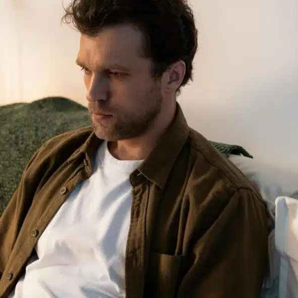 A man sitting on a couch in a softly lit room, appearing deep in thought.