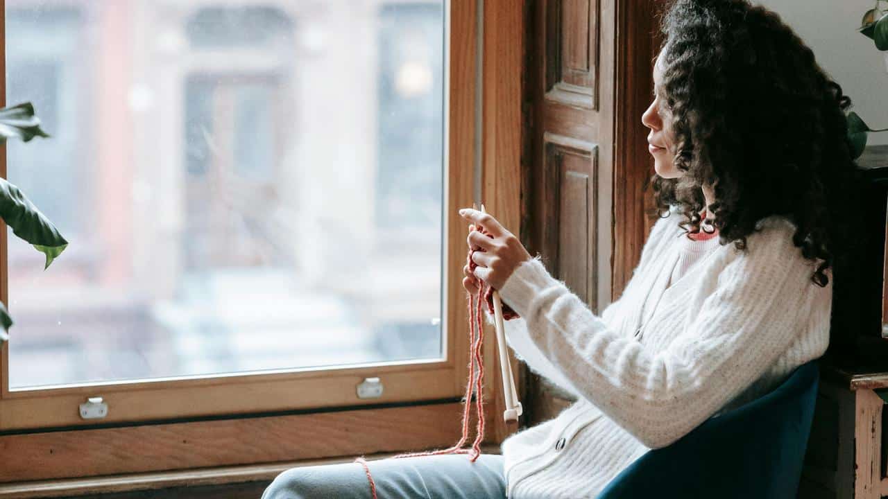 A woman sitting by a window knitting with red yarn.