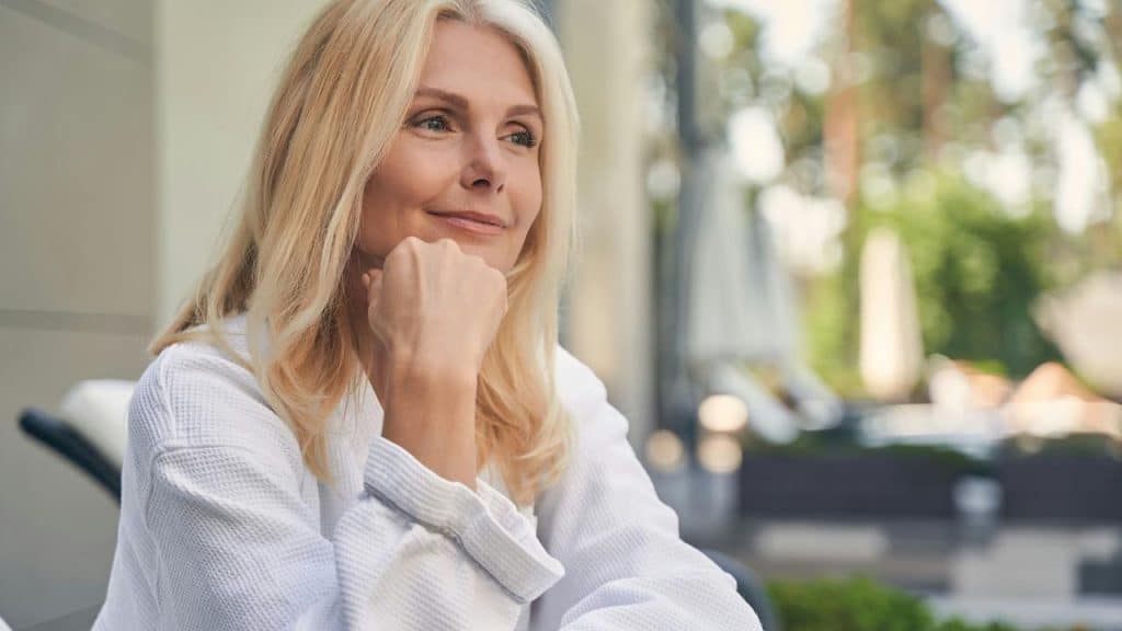 A woman in a white robe sitting outdoors and smiling thoughtfully.