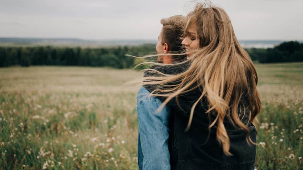 A couple embracing in a grassy field, with the woman’s hair blowing in the wind.