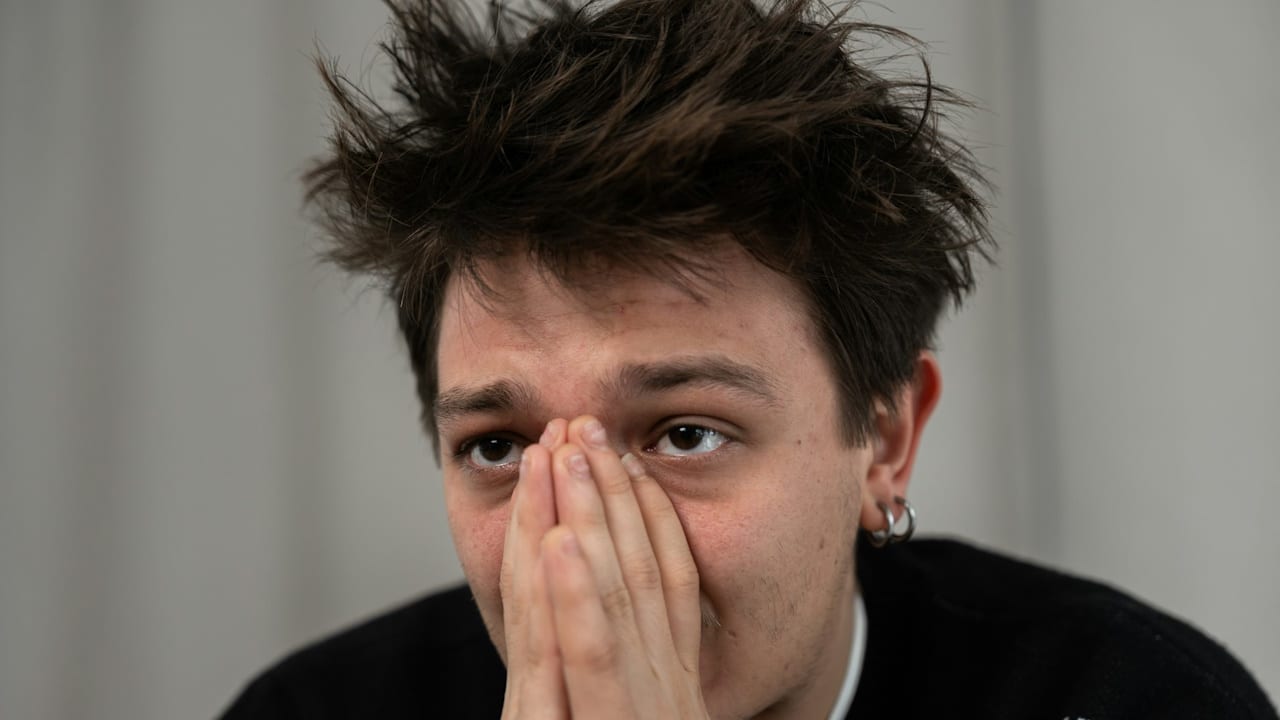 A close-up of a young man with messy hair covering his mouth and nose with his hands, looking worried or anxious.