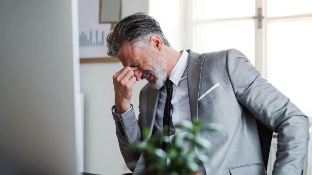 A man in a grey suit sits at a desk, pinching the bridge of his nose.