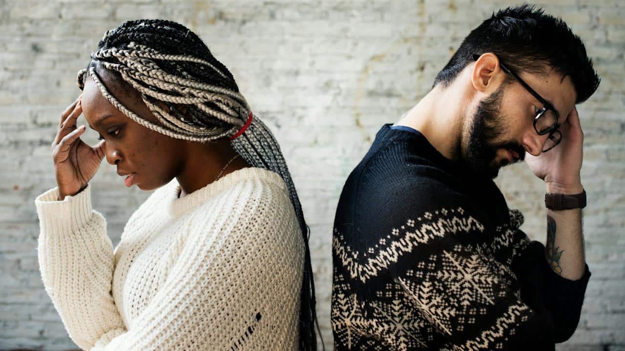 A couple standing with their backs to each other and holding their hands up to their foreheads.