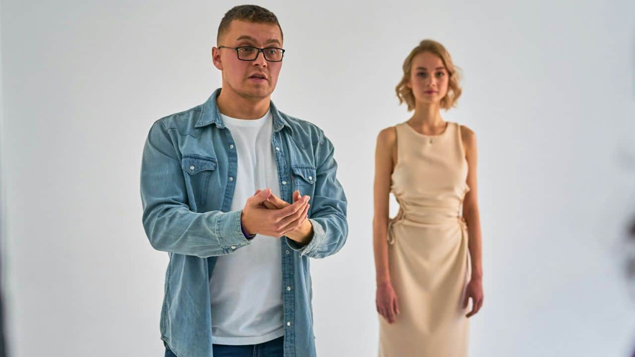 A man in a denim shirt gestures while speaking as a woman stands behind him.