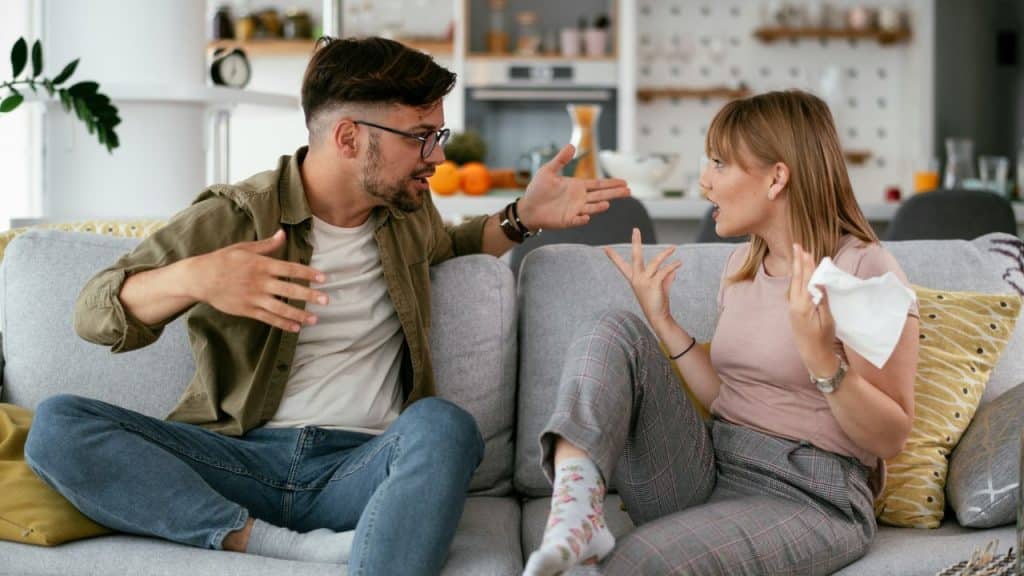 A man and woman sit on a grey sofa, gesturing with their hands while talking.