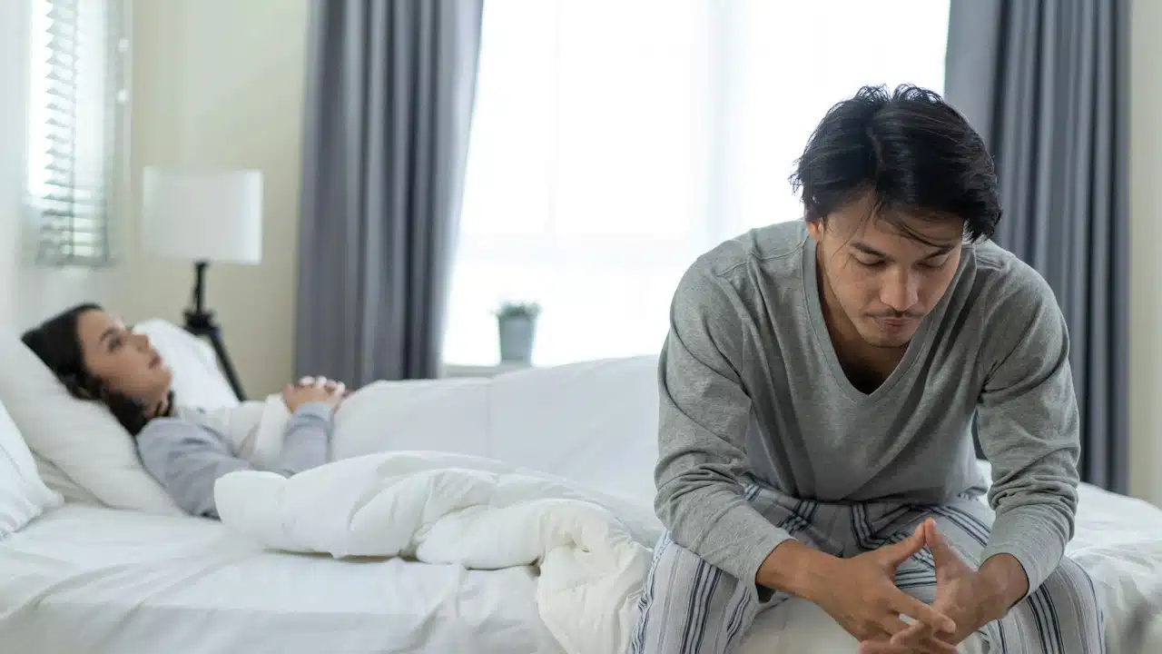 A man sitting on the bed looking anxious and clasping his hands while a woman lies in the background.