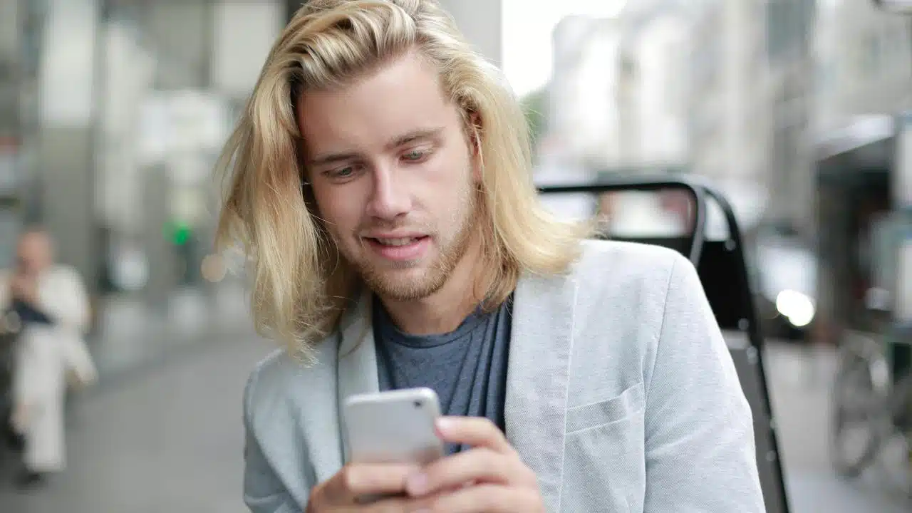 A long-haired man in a light blazer looking at his smartphone while sitting outdoors in a city.