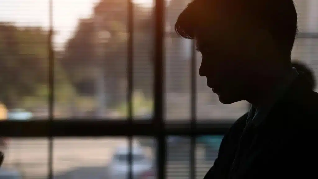 A silhouetted person standing near a window with soft light behind them.