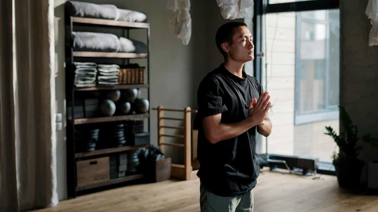 A person standing indoors with hands together, appearing to meditate.
