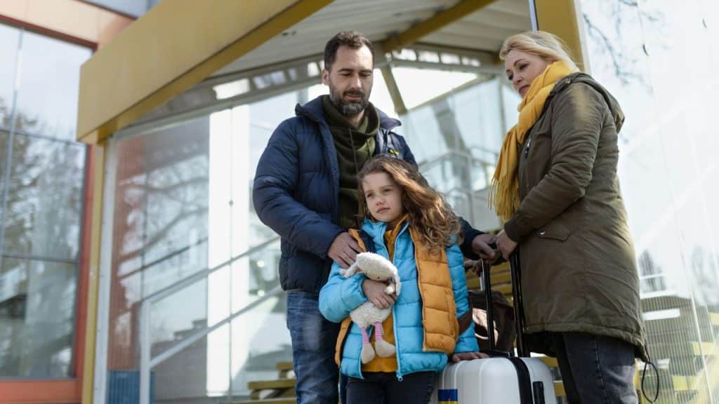 A man, woman, and child stand outside with a suitcase near a glass building.