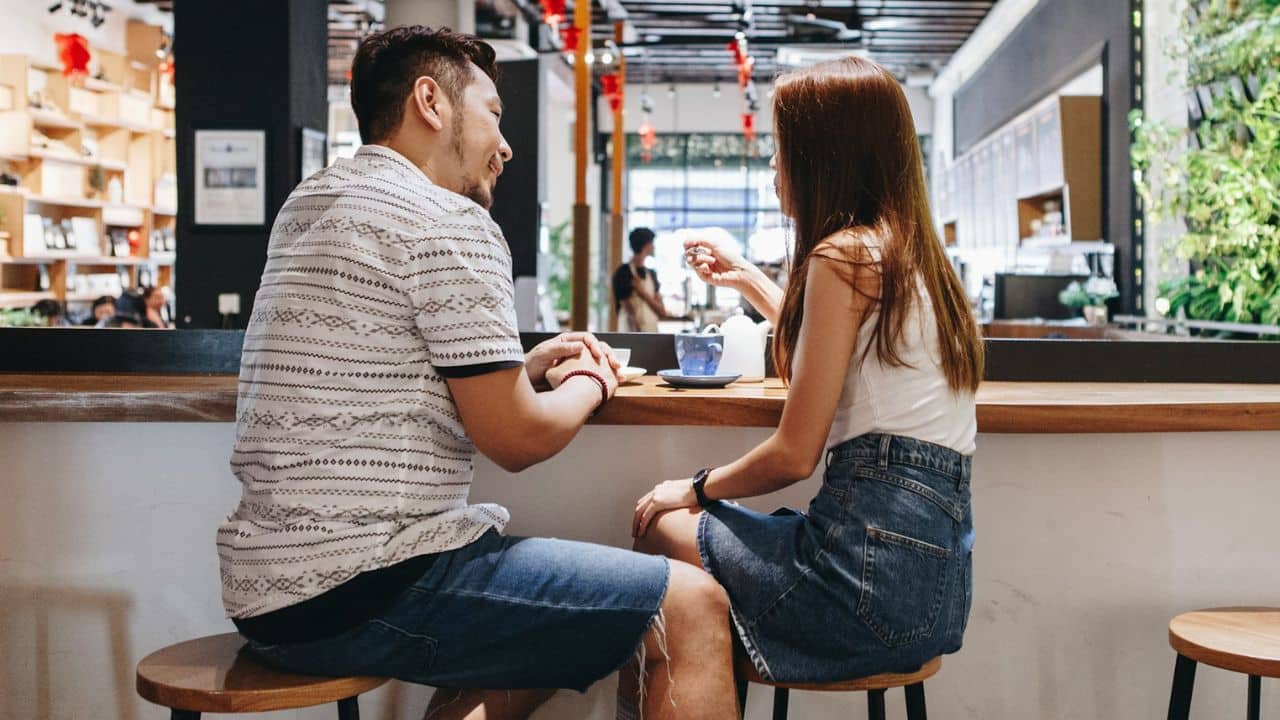 A man and woman sit at a cafe counter talking over coffee and tea.
