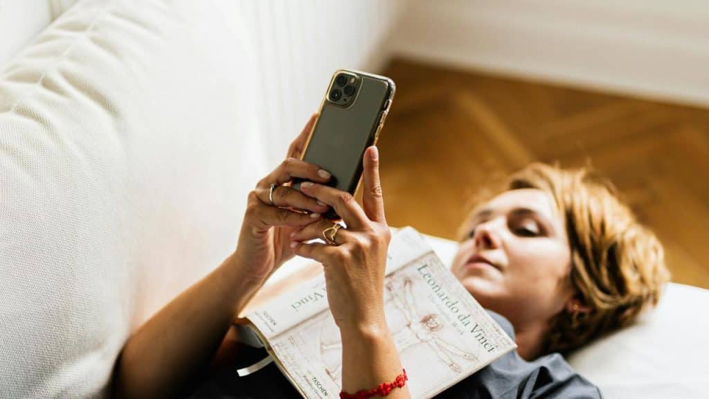 A woman lying on a couch using a smartphone with a book on her chest.