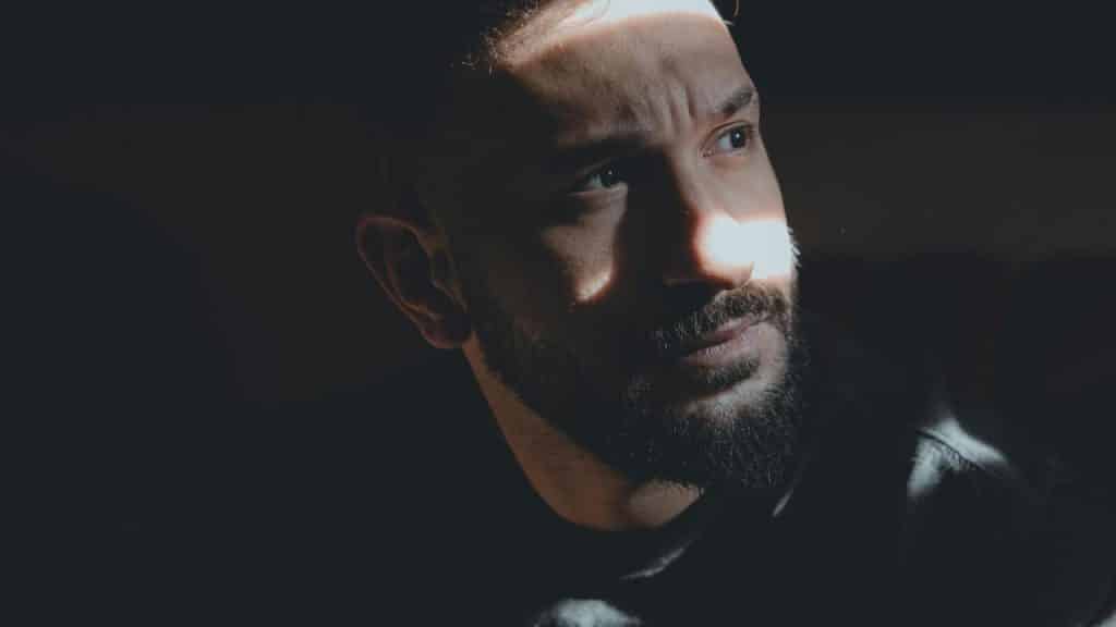 A bearded man looks to the side in dramatic low light.