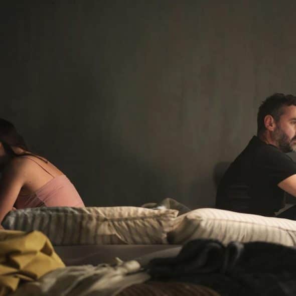 A couple sitting back-to-back on a bed in a dimly lit room.