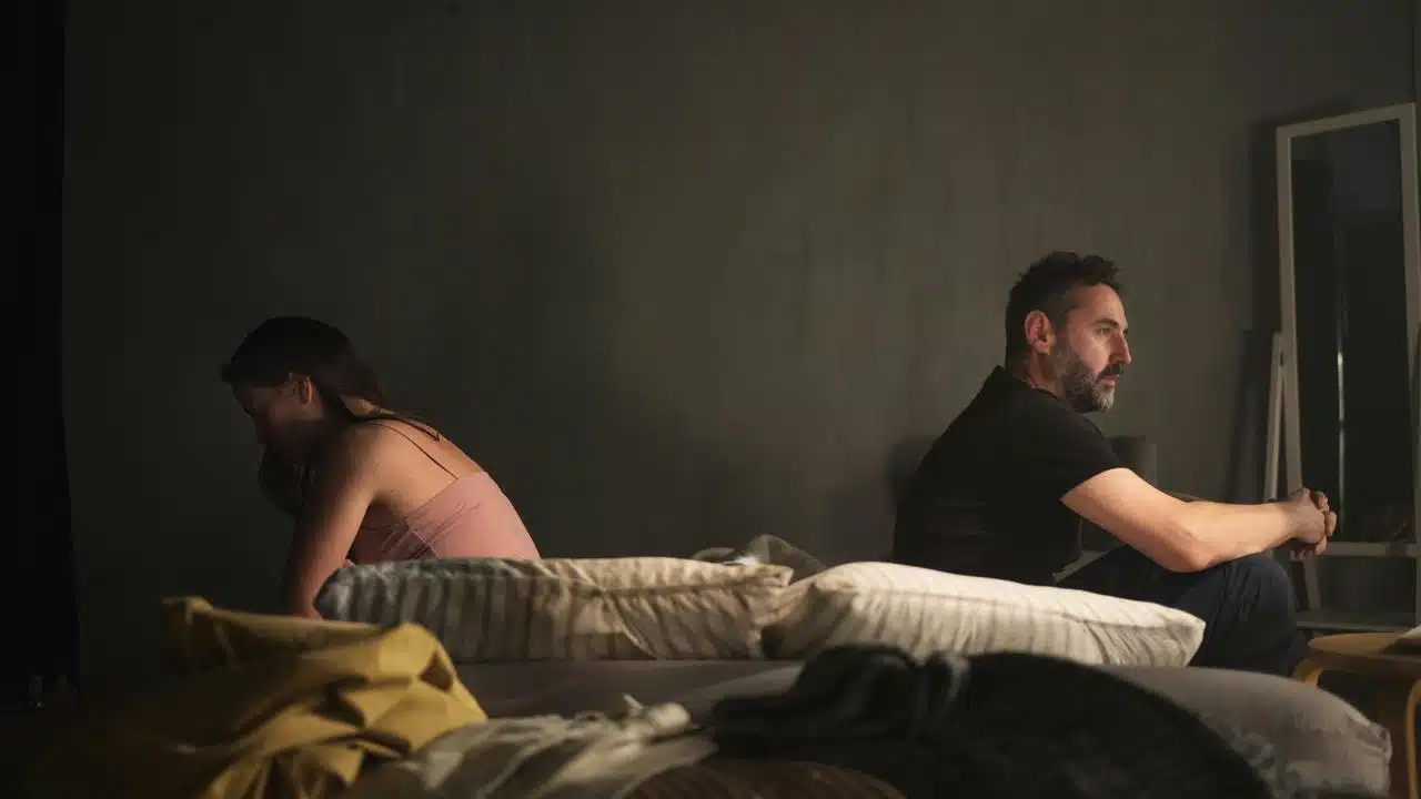 A couple sitting back-to-back on a bed in a dimly lit room.