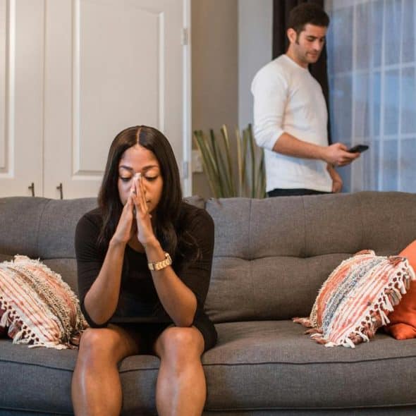A distressed woman on a couch with a man on his phone in the background.