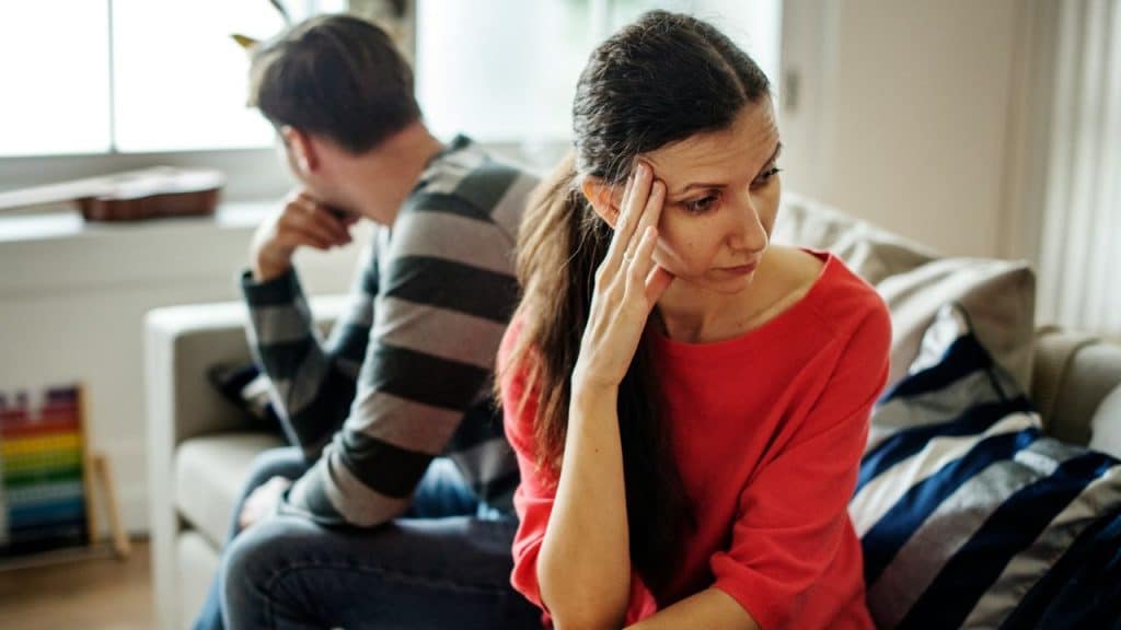 A woman in a red shirt sits on a sofa looking away from a man.