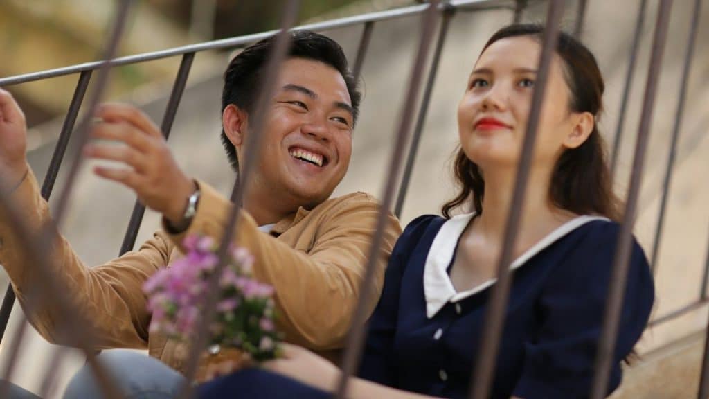A man laughs and holds flowers while a woman next to him looks away.