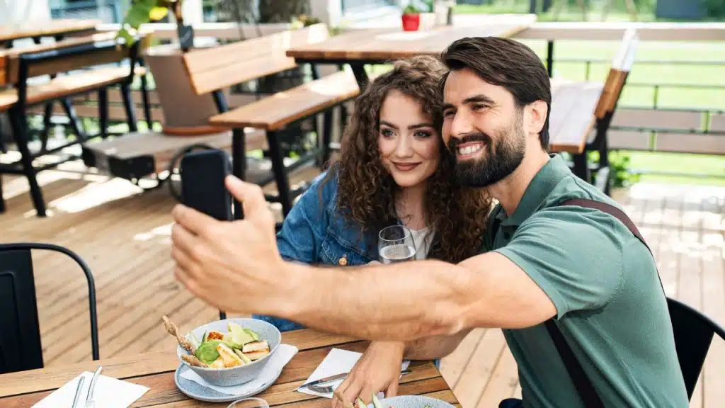 A bearded man holds a smartphone to take a selfie with a woman at lunch.
