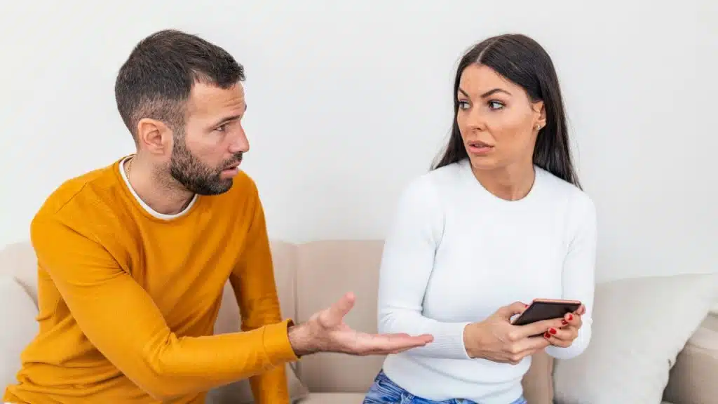 A man in a yellow sweater gestures toward a woman holding a smartphone on a sofa.