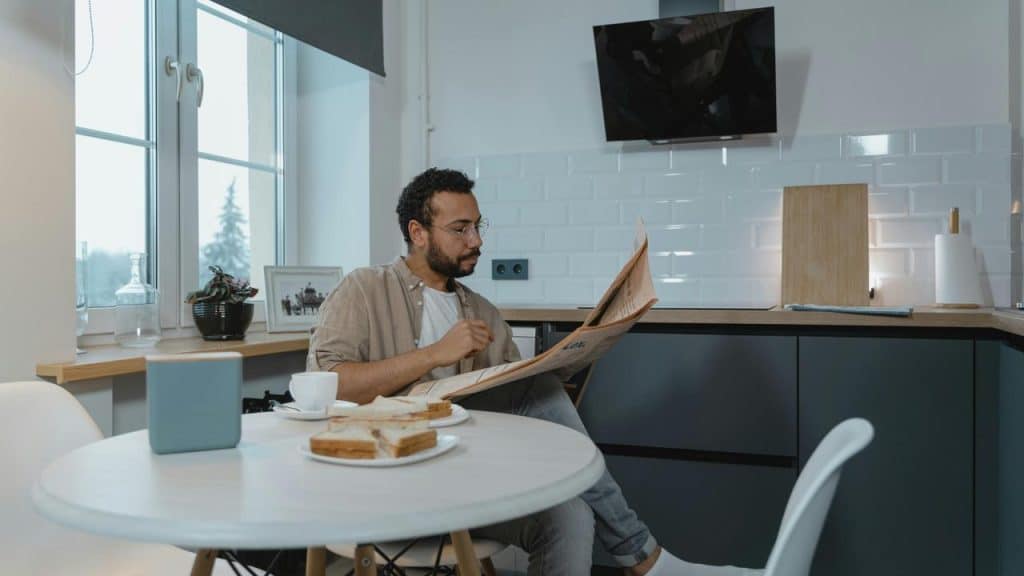 A man reading a newspaper at a kitchen table with coffee and toast.