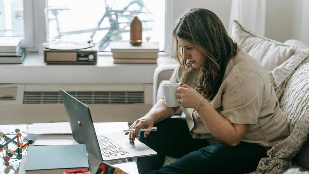 A woman using a laptop while holding a mug and pen on a couch.