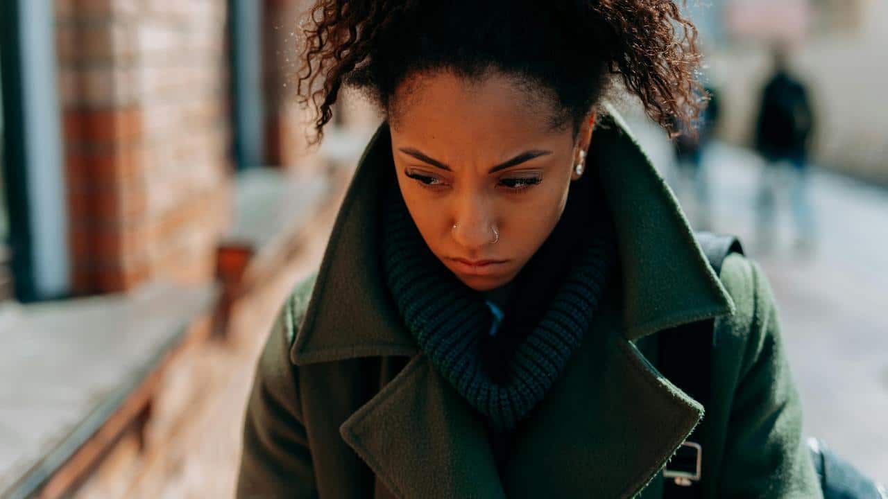 A woman in a green coat looking down with a worried expression on a city street.