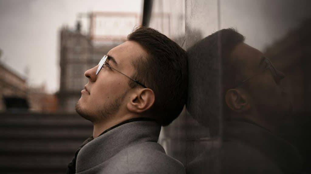 A serious man with glasses and a beard is leaning his head against a stone wall, looking up.