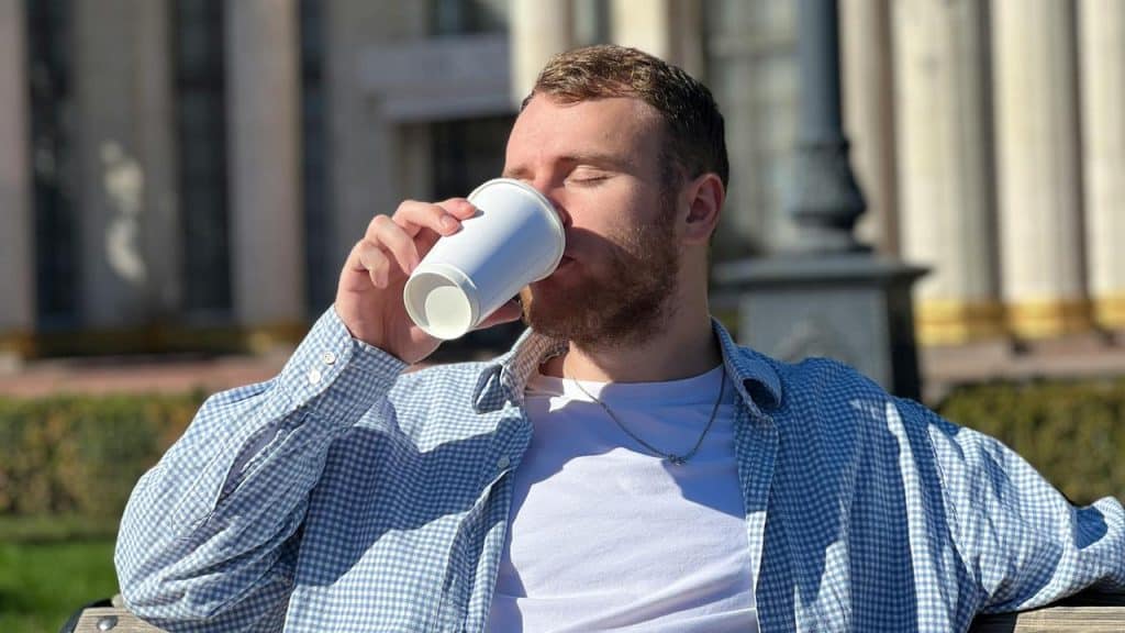 A man sitting outdoors drinking from a takeaway cup with eyes closed.