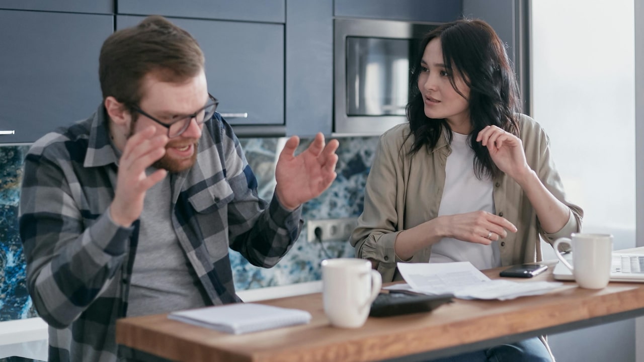 A man and woman sit at a kitchen counter with papers, looking stressed and arguing.