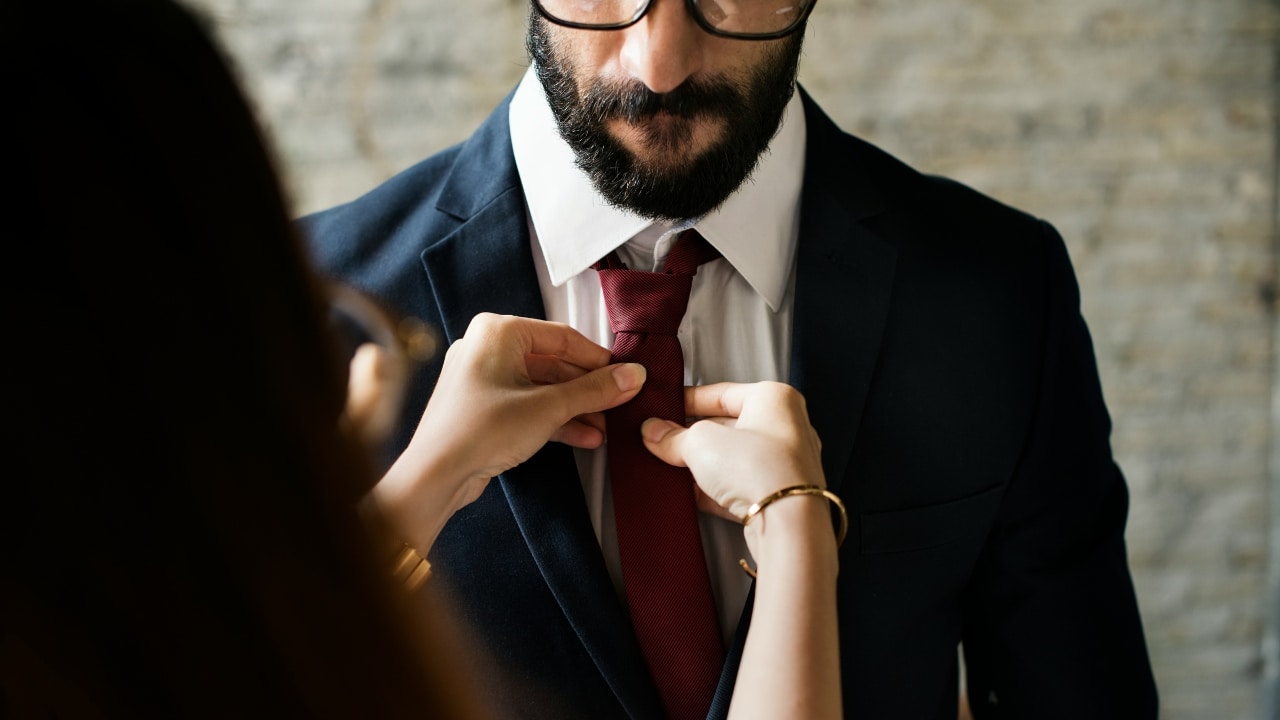 A person's hands adjust a red necktie on a bearded man wearing a dark suit and glasses.