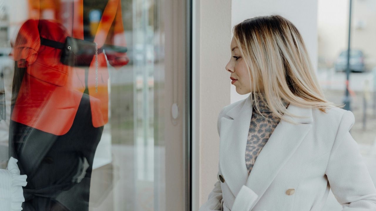 A blonde woman in a white coat looks at a red jacket in a window.