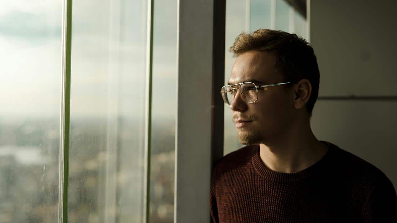 A man wearing glasses gazing out a window in soft daylight.