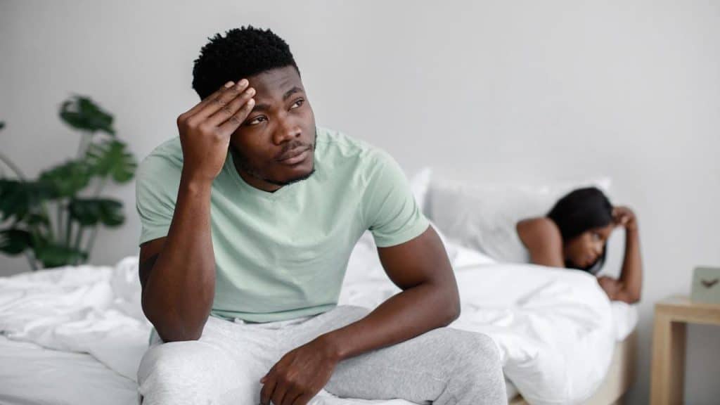 A man sits on a bed looking stressed with his hand on his forehead, while a woman lies facing away from him in the background.