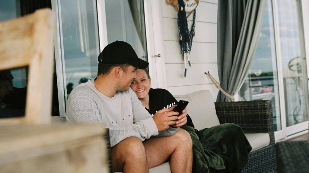 A couple sitting close on a porch, smiling while looking at a phone together.
