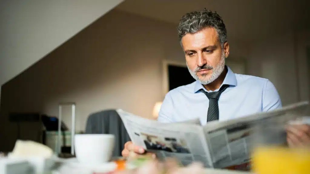 A middle-aged man reading a newspaper at a table with a cup of coffee nearby.