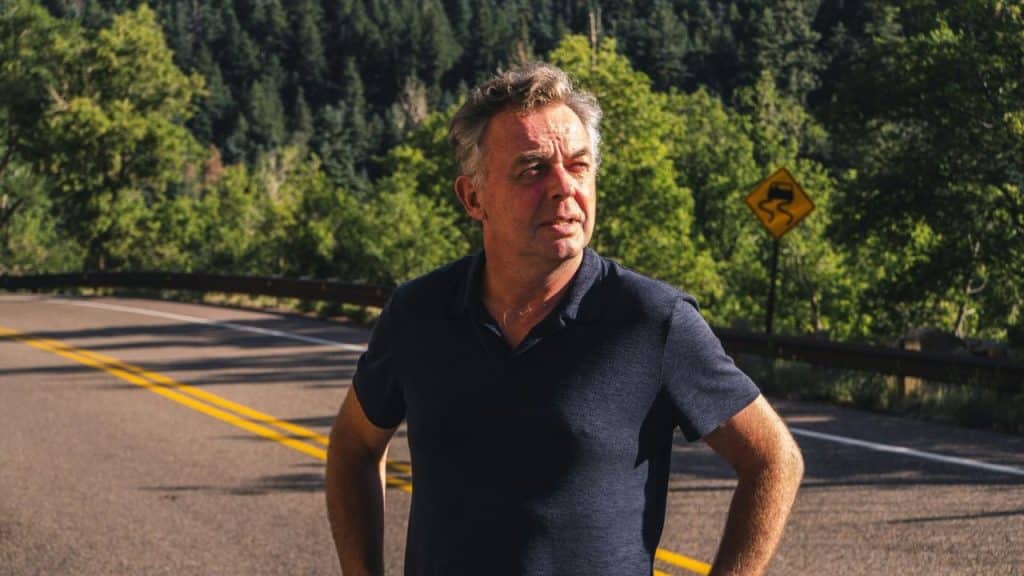 A man in a dark polo shirt stands on a road with trees behind him.