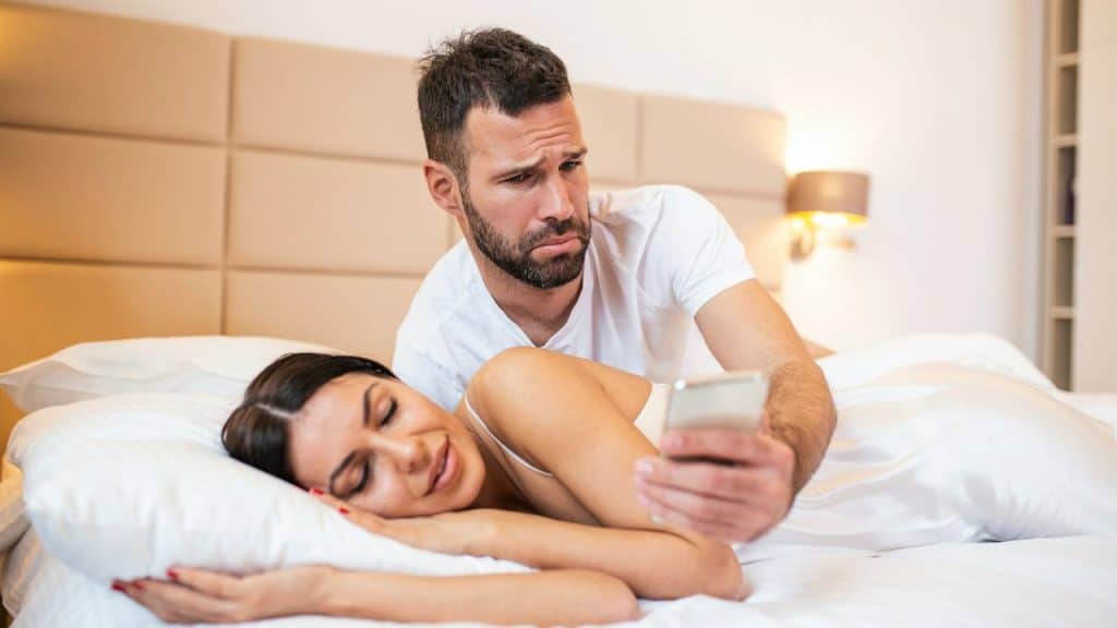 A man looks at a phone while a woman lies asleep in a bed beside him.