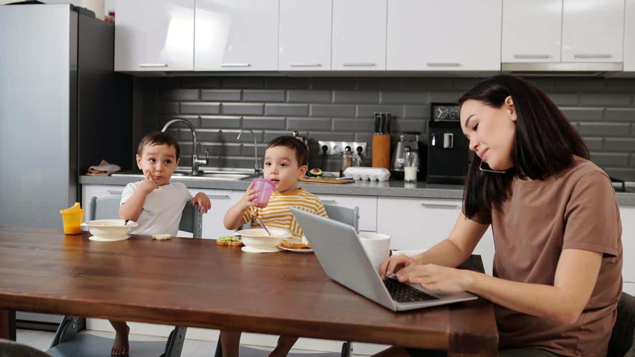 A mother working on a laptop while her children eat breakfast.