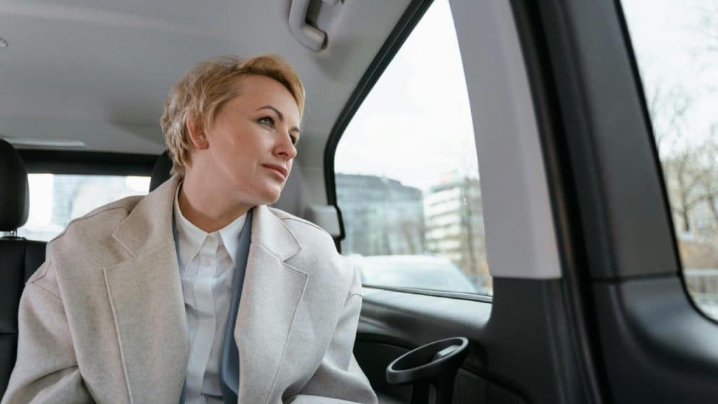 A woman in a light coat looks out the car window thoughtfully.