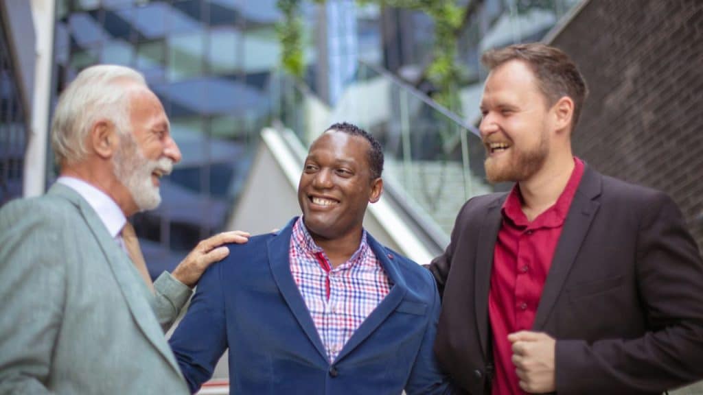 Three men in jackets stand close together outdoors, laughing and smiling with one another.