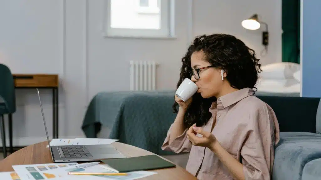 A woman sitting at a table drinking coffee while working on a laptop.