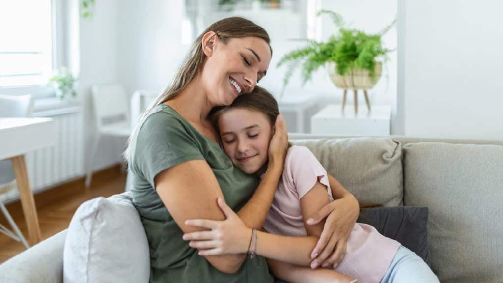 A smiling woman and a young girl sit on a couch sharing a warm embrace.
