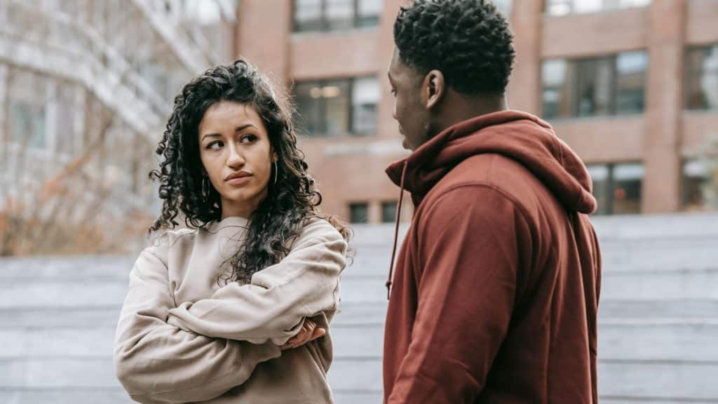 A woman with curly hair and crossed arms looks at a man in a hoodie.