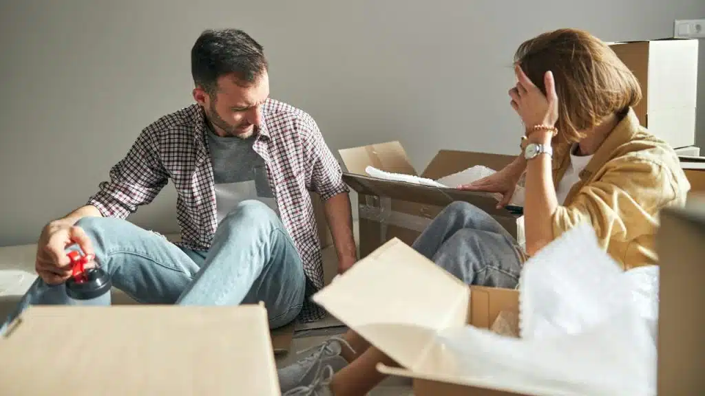 A man and a woman sit on the floor surrounded by open cardboard packing boxes.