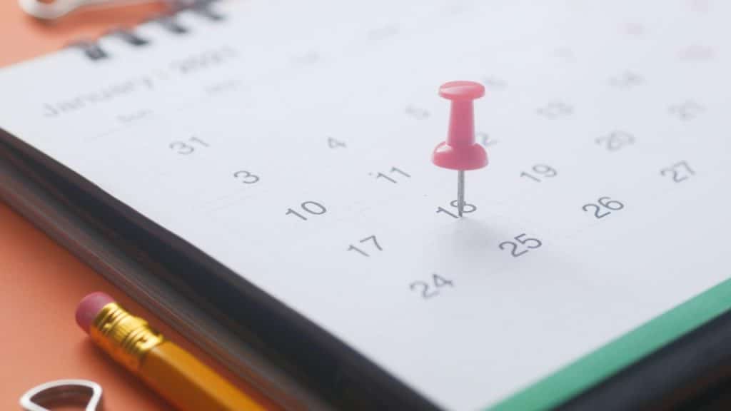 A pink pushpin is stuck into the number eighteen on a white desk calendar.