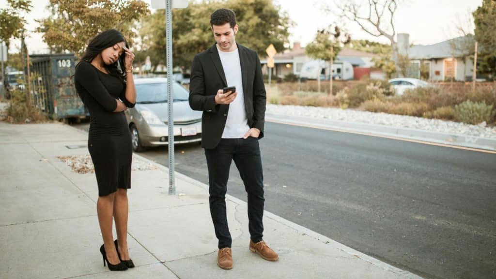 A woman rests her head on her hand while a man looks at his smartphone.