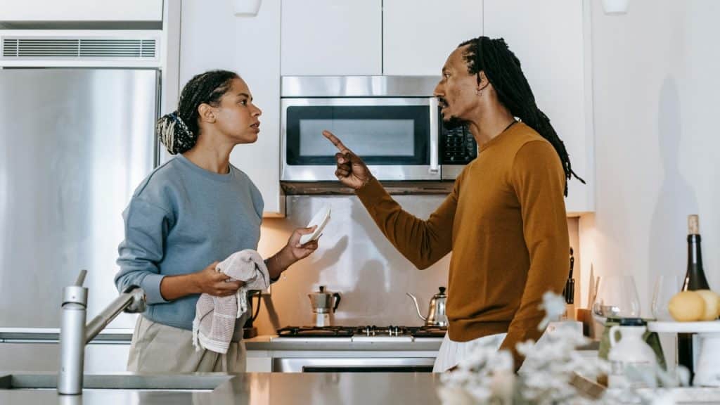 A man points a finger toward a woman holding a white plate in a kitchen.