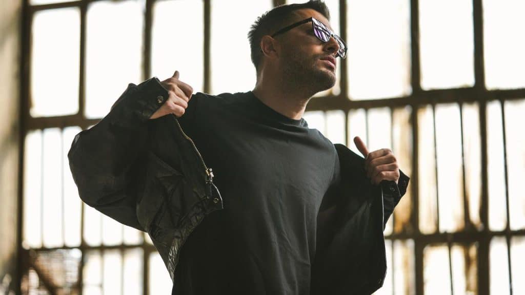 A man wearing sunglasses putting on a jacket indoors.
