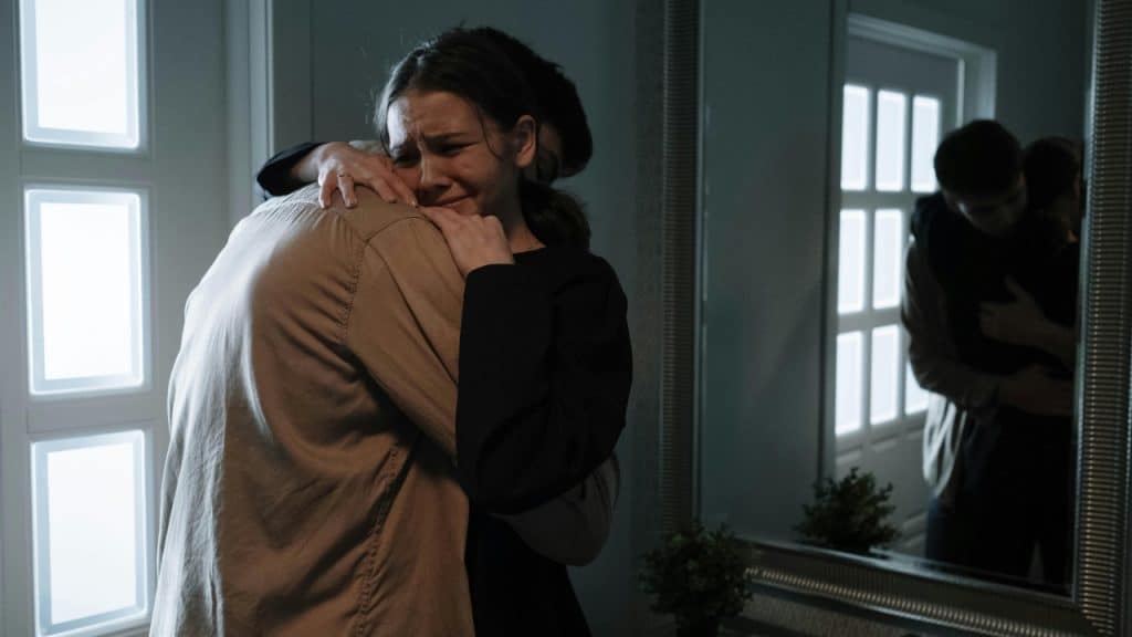 A woman cries while hugging a man in a dimly lit room near a large mirror.